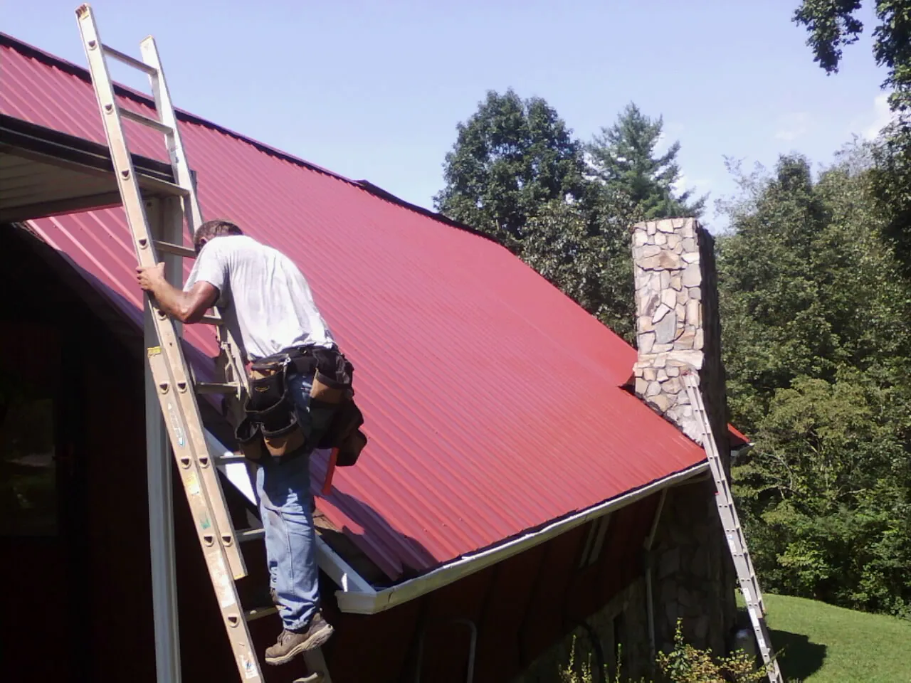 Roofer on a ladder beside a red metal roof and stone chimney — Soffit & Fascia Repair by Roofing Brothers in Miamiville